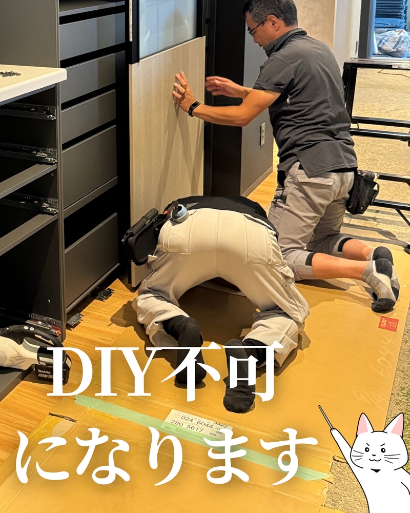 黒いキッチン収納キャビネントの横で、専門業者2名が2wayクローゼットの設置作業を行っている様子。1人はしゃがんでパネルを支え、もう1人は膝をついて床の梱包材を確認している。床には養生シートと梱包用段ボールが敷かれている。画像には「DIY不可になります」の白文字と、指差しポーズの猫イラストが添えられている。