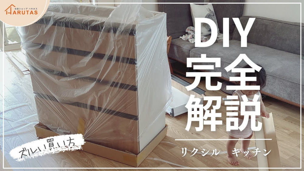 住設ショップHARUTASのロゴ付き画像。梱包用ビニールに包まれたリクシルキッチンの引き出しユニットが室内に置かれ、その奥でグレーのソファ前の床に子供が座っている。右側に大きな白文字で「DIY完全解説」、その下に「リクシル キッチン」の文字。左下には「ズルい買い方」という手書き風の文字が添えられている。