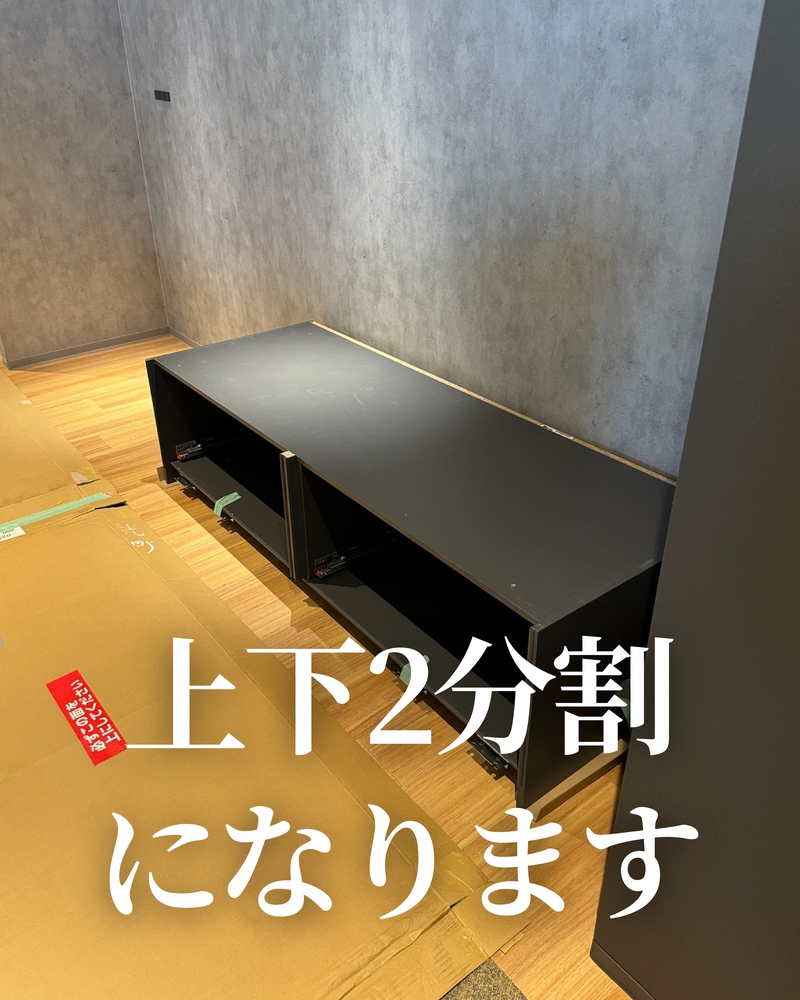 設置現場の写真。グレーの壁と木目調の床の部屋に、黒色のリシェル2wayクローゼットのキャビネット下部が仮置きされている。引き出しレールが見え、周囲にはダンボールの緩衝材が敷かれている。画像下部に大きな白文字で「上下2分割になります」と記載され、配送時にキャビネットが上下2つに分かれることを示している。