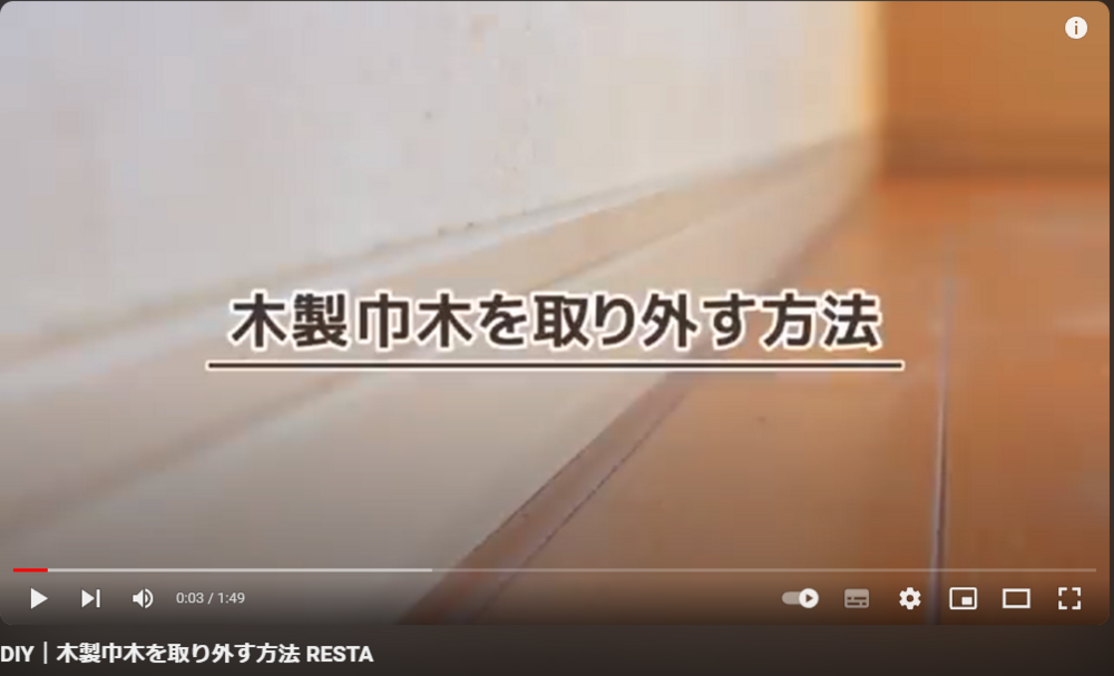 YouTube動画の再生画面。壁と床の境目にある白い木製巾木がアップで映り、「木製巾木を取り外す方法」と大きな文字で表示されている。再生時間は0:03／1:49。下部にタイトル「DIY｜木製巾木を取り外す方法 RESTA」と表示され、再生バーや音量、字幕、設定などの操作アイコンが並ぶ。