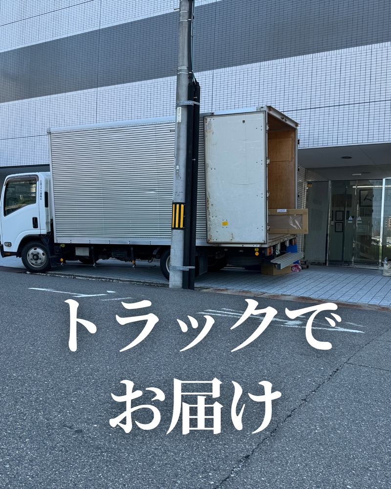 ビルの入口前に停車した白い平ボディトラックの荷台後部が開き、木製の梱包箱が積まれている配送風景の写真。タイル張りの建物とガラス扉の入口が背景に見える。画像下部に大きな白文字で「トラックでお届け」と記載されており、2wayクローゼットのキャビネットがトラックで配送される様子を示している。