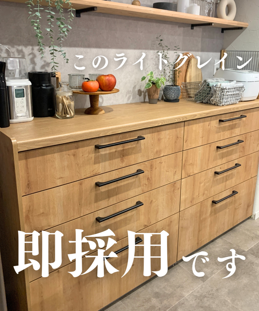 木目調カウンター「ライトグレイン」の引き出し付きキッチンカップボード。黒いバー状の取っ手が並び、天板上にはコーヒーメーカー、瓶、柿とりんごを乗せた木製スタンド、観葉植物、まな板などが置かれている。文字で「このライトグレイン」「即採用です」と記載。