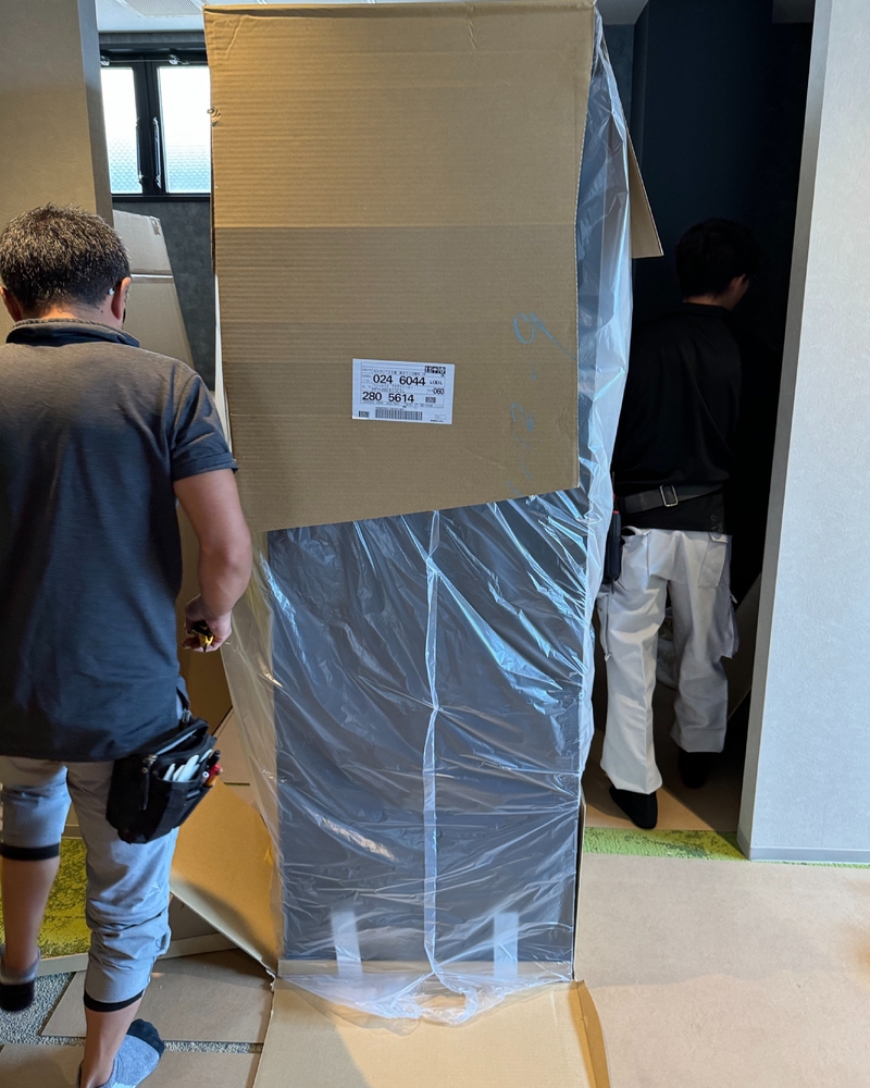 段ボールと透明ビニールに梱包された2wayクローゼットのキャビネットを、業者2名が室内へ搬入している様子。梱包箱には「024 6044」「280 5614」と記載されたラベルが貼付されている。作業員は工具ベルトを装着し、上下2分割された大型キャビネットを慎重に運んでいる。