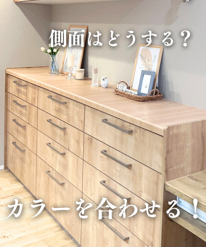 木目調カウンターと引き出しが並ぶキッチンカップボードの写真。天板の上には花瓶、キャンドル、フォトフレーム、フクロウの置物、ウェルカムと書かれたかごが飾られている。画像上部に「側面はどうする？」、下部に「カラーを合わせる！」という白文字が配置され、側面カラーを本体と統一するポイントを示している。