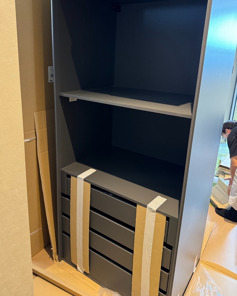 設置作業中の2wayクローゼット本体。上部は開放的な棚板付き収納スペース、下部は複数段の引き出しで構成され、引き出し前面は保護のため段ボールとテープで固定されている。奥では作業員が搬入・設置作業を行っている様子が見える。
