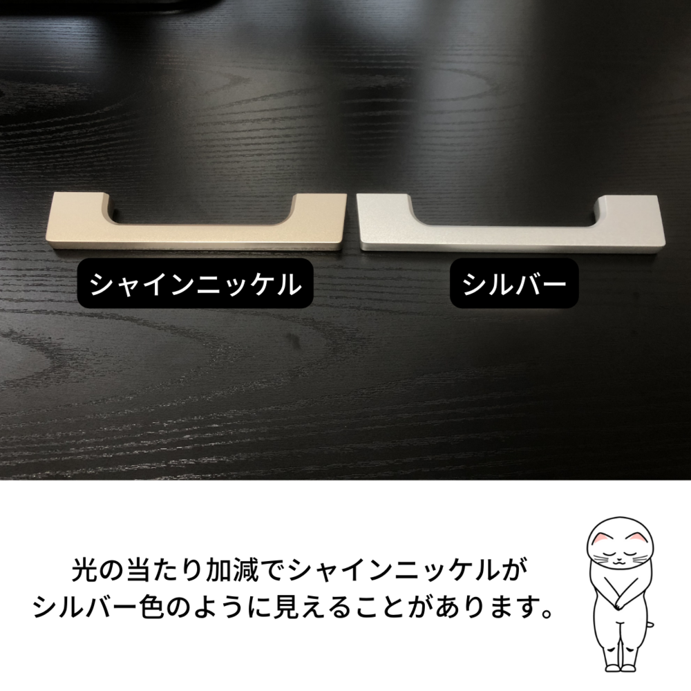 黒い木目調のテーブルの上にコの字型の取っ手が2つ並べて置かれた写真。左側には「シャインニッケル」、右側には「シルバー」とラベルが付いている。両者は色味が非常に似ており見分けにくい。下部には「光の当たり加減でシャインニッケルがシルバー色のように見えることがあります。」という説明文と、目を閉じたイラストの猫キャラクターが添えられている。