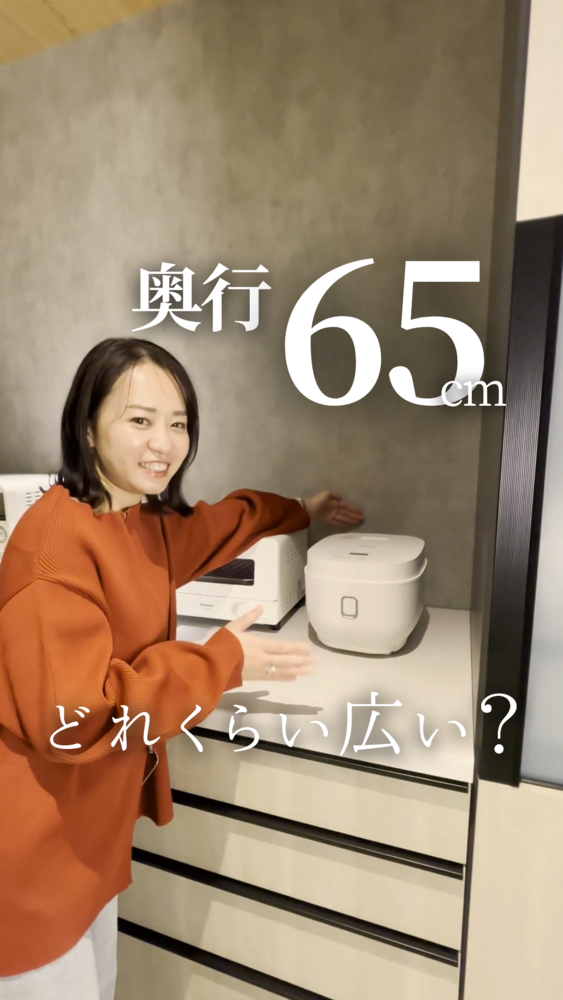 オレンジ色のニットを着た女性が笑顔でキッチン収納カウンターを手で示している写真。カウンター上にはトースターと白い炊飯器が置かれている。画像上部に「奥行65cm」、下部に「どれくらい広い？」の文字が大きく表示されており、リシェルの奥行65cmキャビネットの実際の広さを紹介している場面。