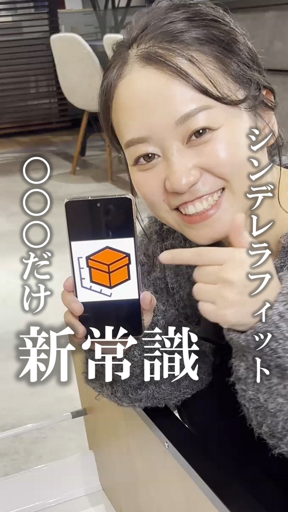 笑顔の女性がスマートフォンを掲げて画面のオレンジ色の収納ボックスのアイコンを指さしている写真。背景にはショールームの椅子やテーブルが見える。画像には「シンデレラフィット」「〇〇〇だけ新常識」という文字が縦書き・大きな白文字で配置され、収納サイズ選びに関する内容を示すインスタグラム投稿風の画像。