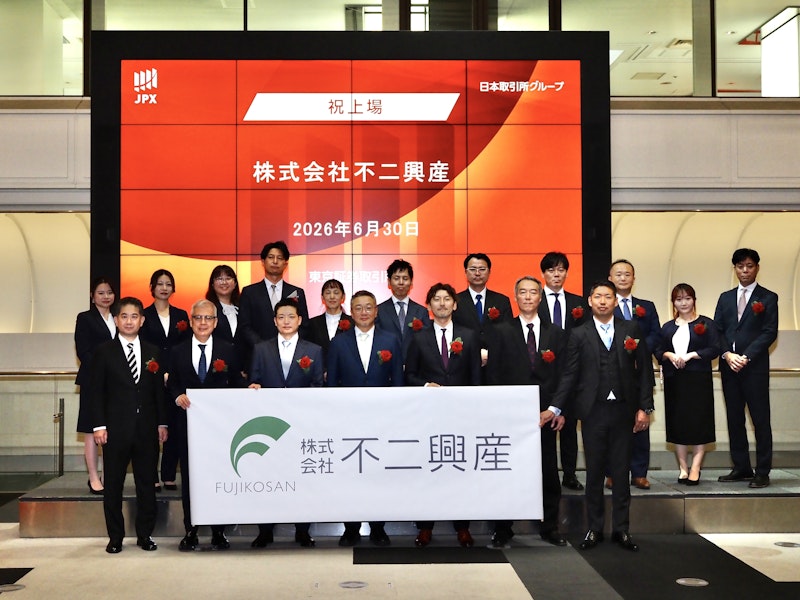 東京証券取引所での株式会社不二興産の新規上場セレモニーの様子。JPXのロゴと「祝上場 株式会社不二興産」と書かれた赤い大画面の前に、スーツ姿の関係者たちが並び、会社ロゴ入りの横断幕を掲げて記念撮影をしている。