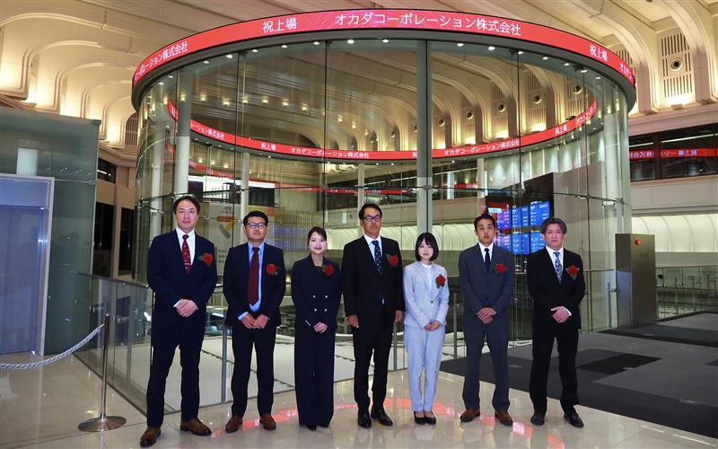 東京証券取引所での上場セレモニーにて、チッカー(電光掲示板)を背に並ぶオカダコーポレーション役職員一同の記念写真