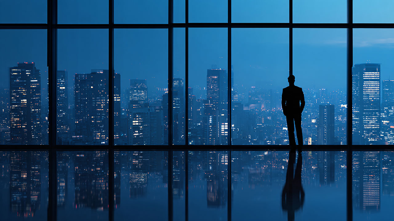 Silhouette of a businessman looking out of a large floor-to-ceiling window at a sprawling city skyline at night, with the blue-toned city lights reflecting clearly on the polished office floor.