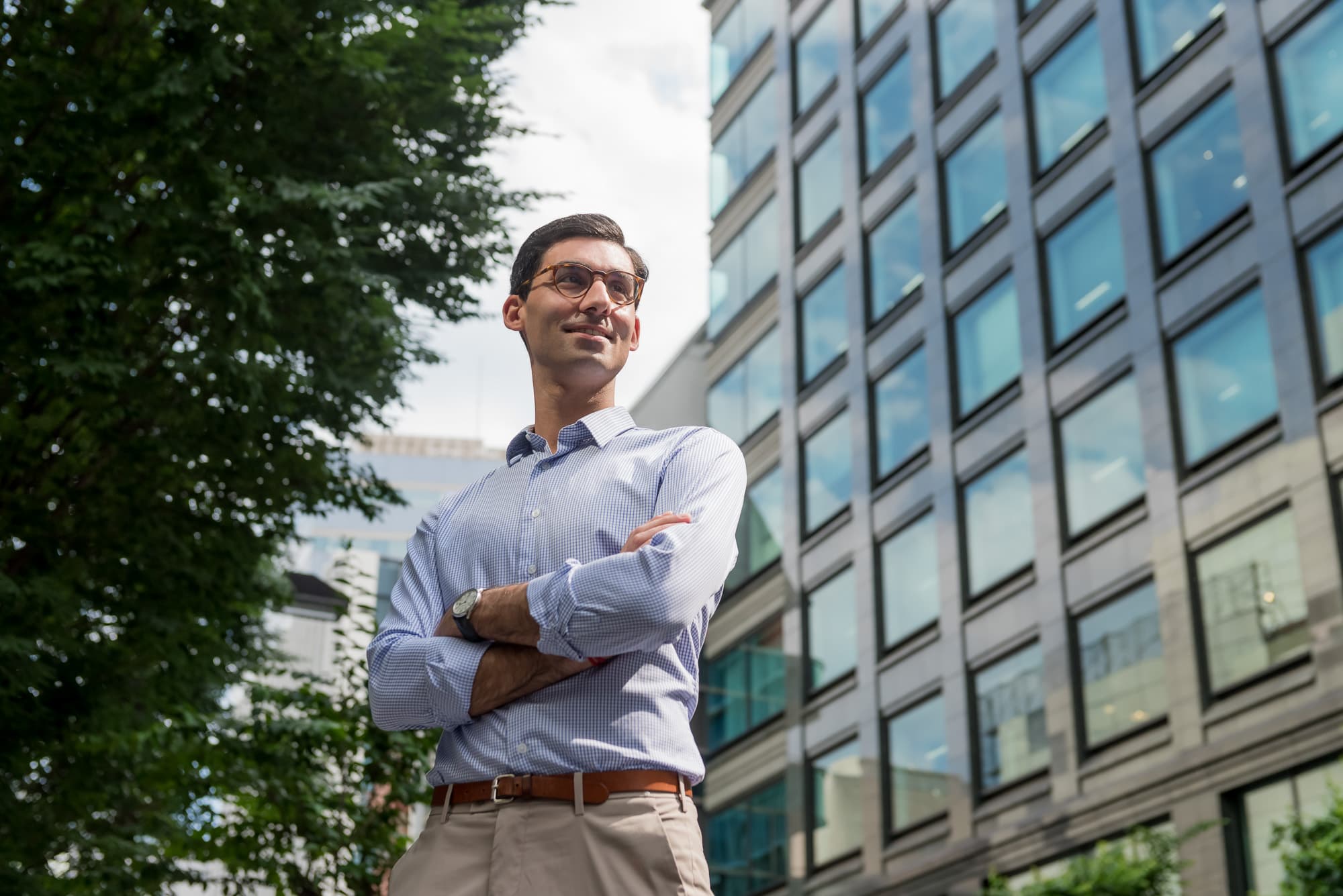 Man in glasses and a dress shirt standing with crossed arms outside a glass building.