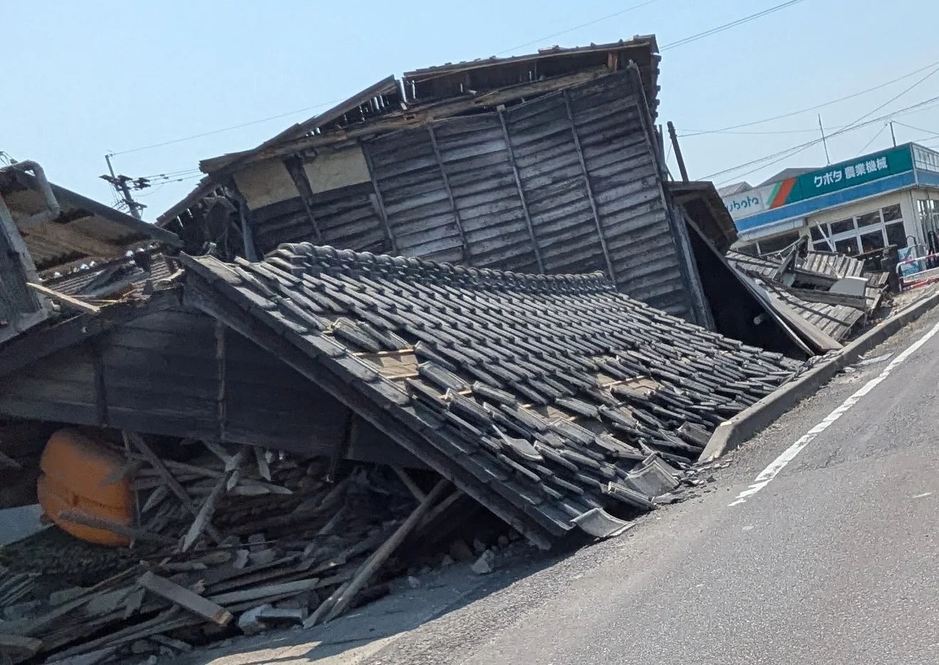 道路沿いで倒壊した木造家屋。屋根瓦が路上近くまで崩れ落ち、奥には外から見て被害のわからない店舗が建っている