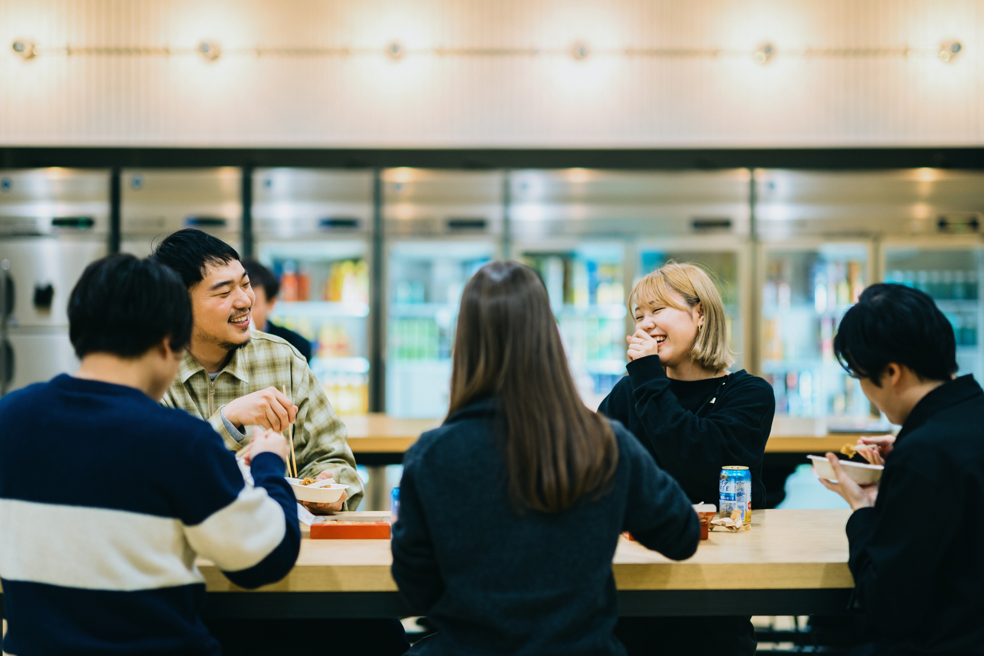 壁一面の冷蔵庫ショーケースの前で飲食しながら談笑する5名の社員の写真