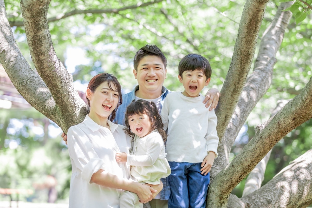 二人の子供と夫婦が家族で寄り添いながら木の下で笑い合っている様子