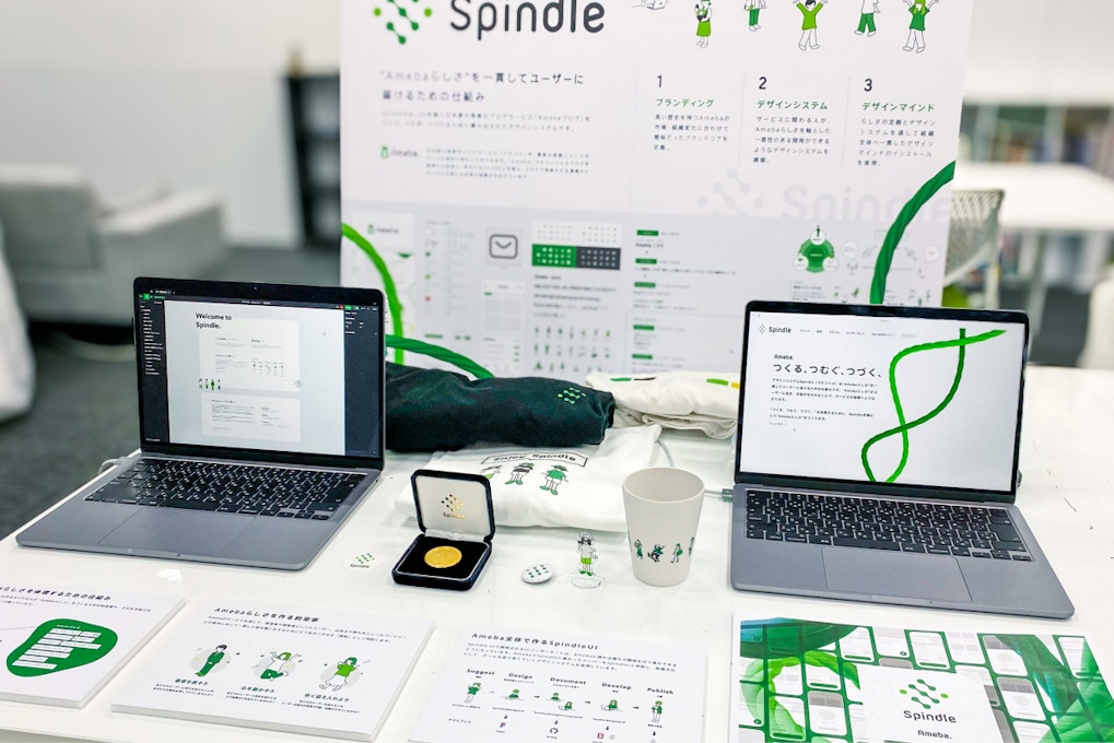 オフィスの一角でグッドデザイン賞の展示をシミュレートしている画像。・Spindleサイト ・Figmaで作成したSpindleのUIライブラリ ・Spindleの概要と実績を説明したドキュメントやパネル ・Spindleのグッズが並んでいる。