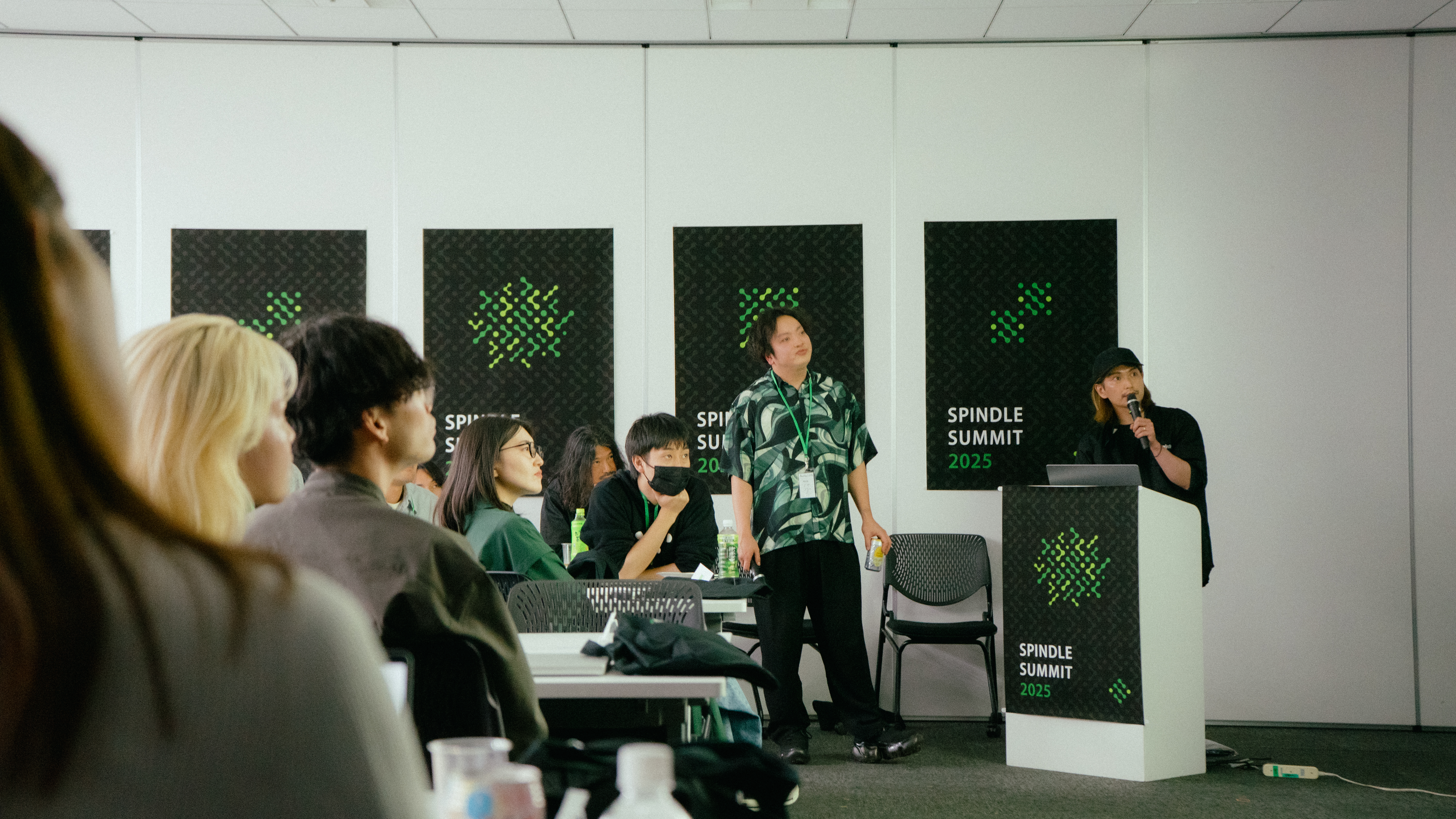 2人の男性がSpindle MCPサーバーについてプレゼンしている様子。ひとりがマイクを持ち、もうひとりはハイボールを持ちスクリーンに目を向けている。