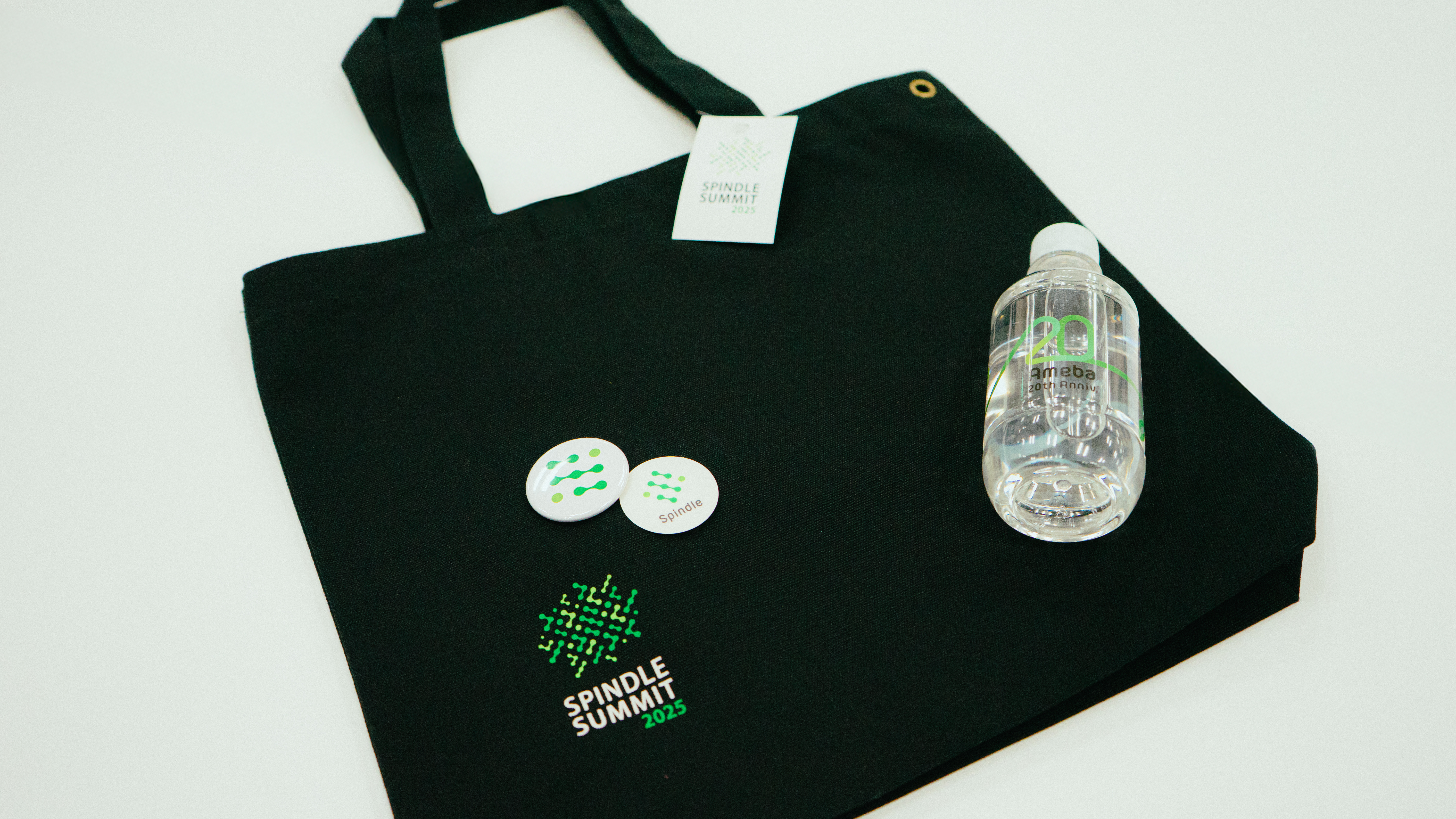 Spindle SUMMIT 2025の参加者向け記念グッズ。黒いトートバッグに加え、ロゴ入り缶バッジと「20周年」記念のミネラルウォーターが並べられている。