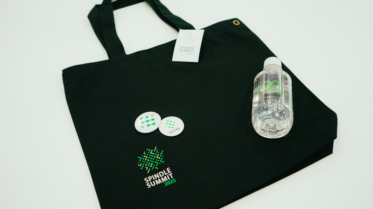 Spindle SUMMIT 2025の参加者向け記念グッズ。黒いトートバッグに加え、ロゴ入り缶バッジと「20周年」記念のミネラルウォーターが並べられている。