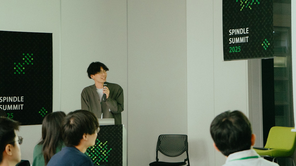 若い男性がスピーカーとして登壇しているシーン。手にマイクを持ち、笑顔で話している姿。