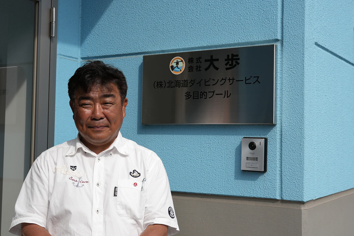 株式会社大歩の社名看板の前に立つ中村さん