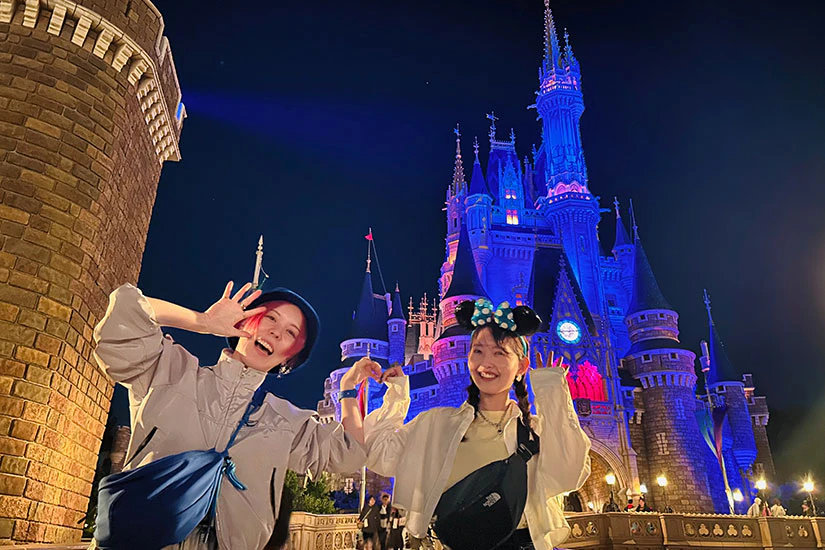 夜のシンデレラ城の前で肘をくっつけてハートをつくる二人のノベルティメンバー