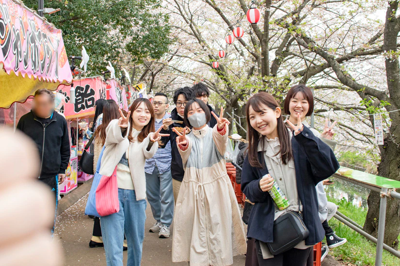 花見海老川の屋台の前で笑顔でポーズを撮るノベルティスタッフの写真を楽しむノベルティメンバー