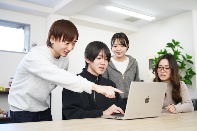 4人のメンバーがテーブルを囲み、MacBookの画面を一緒に確認している様子。