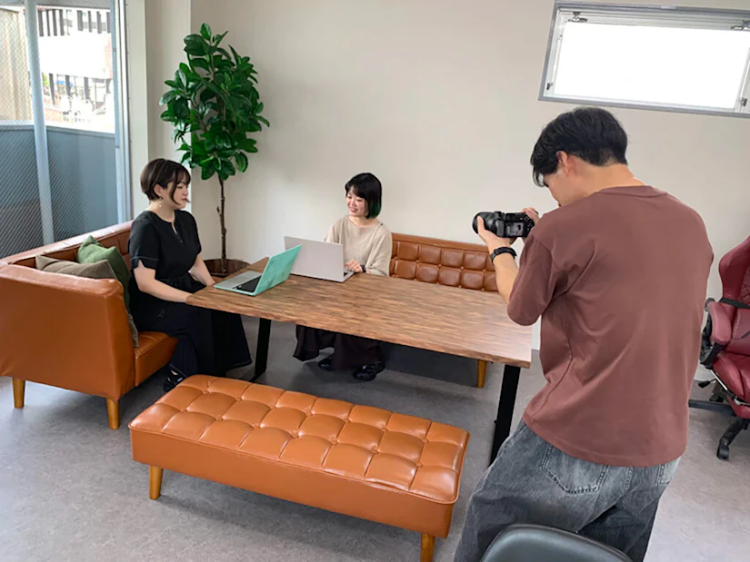 ソファに座り談笑しているノベルティ社員2名とその写真を撮るカメラマン