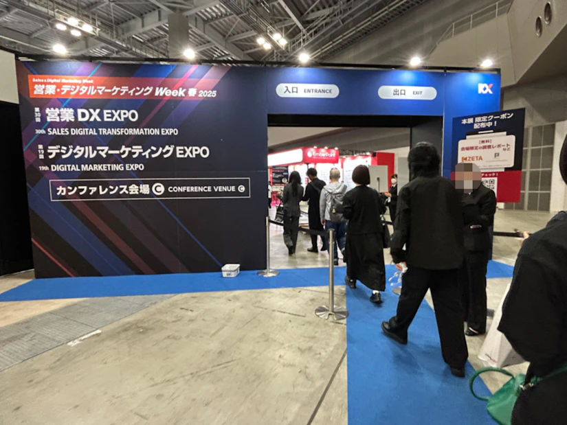 「営業・デジタルマーケティングWeek 春 2025」と書かれたJapan IT Weekの入場ゲートをノベルティ社員がくぐり、会場に入場していく。