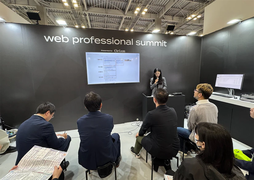 「web professional summit」と書かれたブース中央の登壇スペースにノベルティの社長がマイクを持って立っている。その前にセミナーを聞きにきた来場者が8名ほど、椅子に座ってセミナーを聞いている。