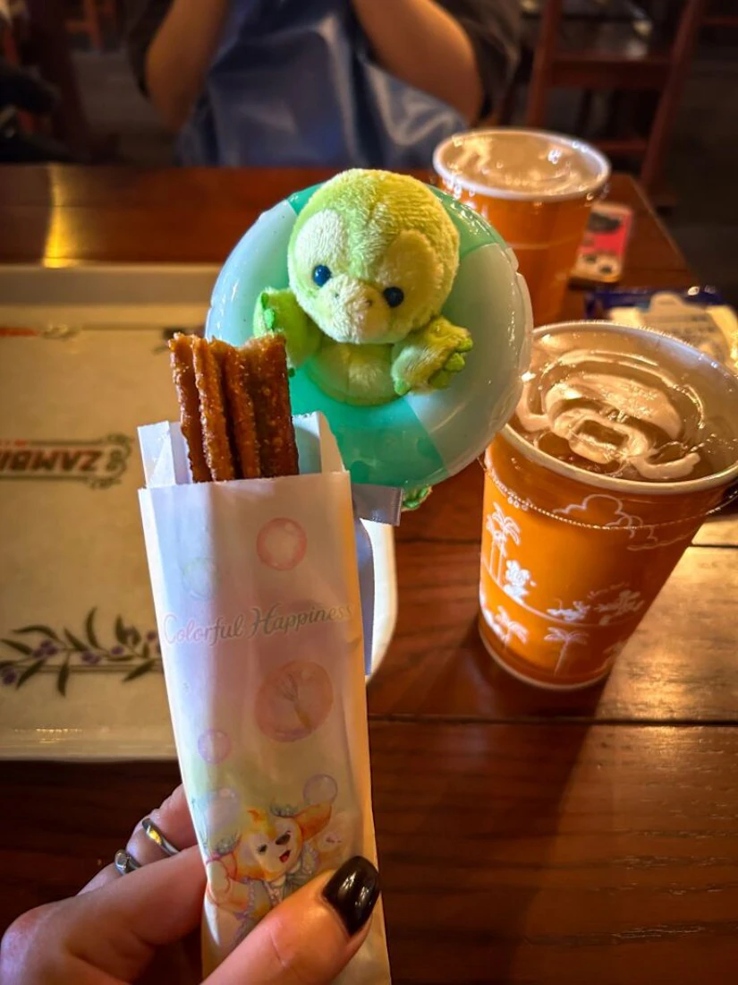 園内で購入したグッズとチュロスの写真。グッズは人気キャラクター「オル・メル」のぬいぐるみスティック。期間限定フレーバーのチュロスと、飲み物と一緒に映っている。