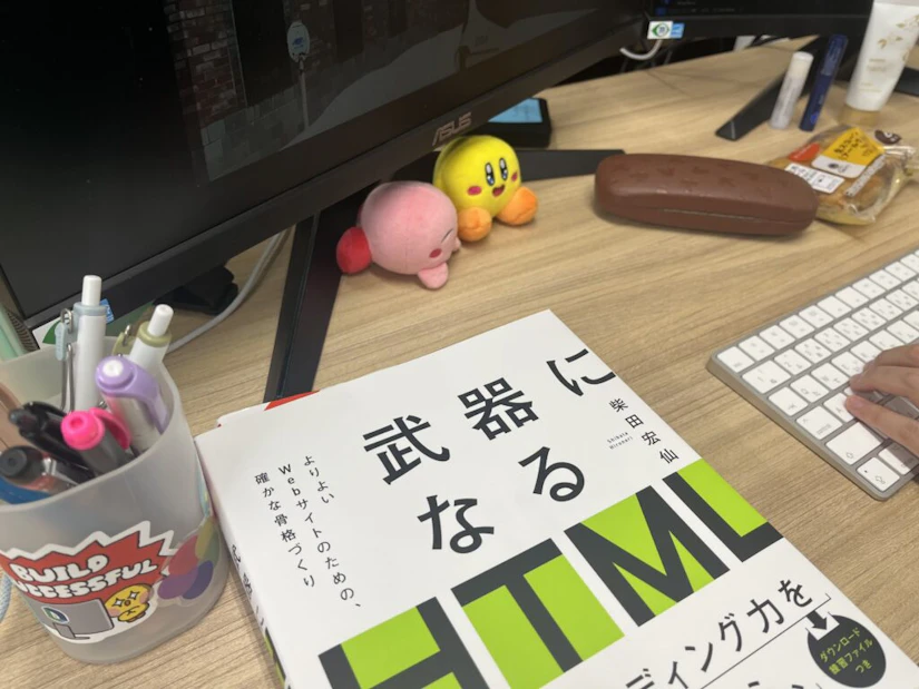パソコンデスク。「武器になるHTML」という本と、ペン立てやメガネケースが置かれている。ピンクと黄色のカービィのぬいぐるみが真ん中に置かれている。