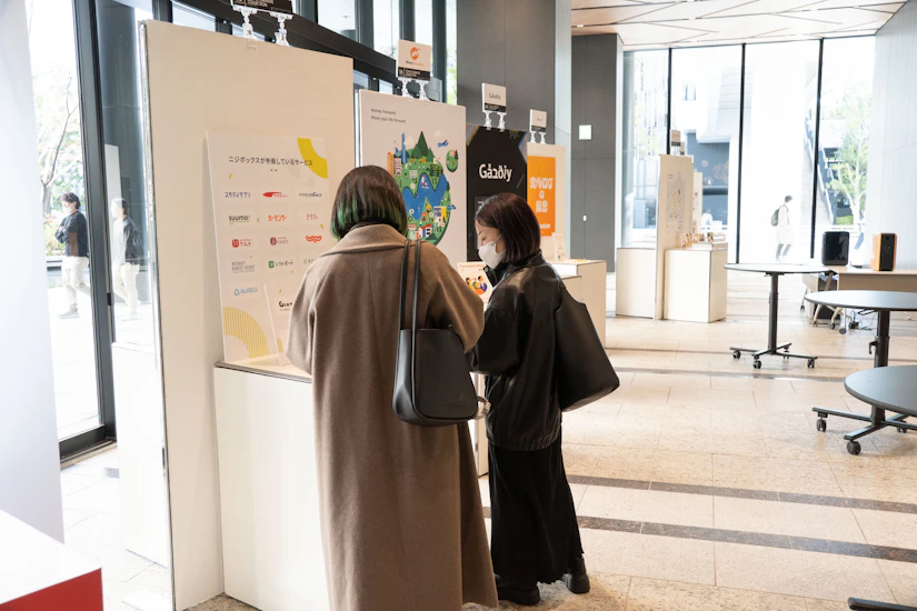 会場の企業ブースで2人の参加メンバーがパンフレットなどのノベルティを眺めている写真