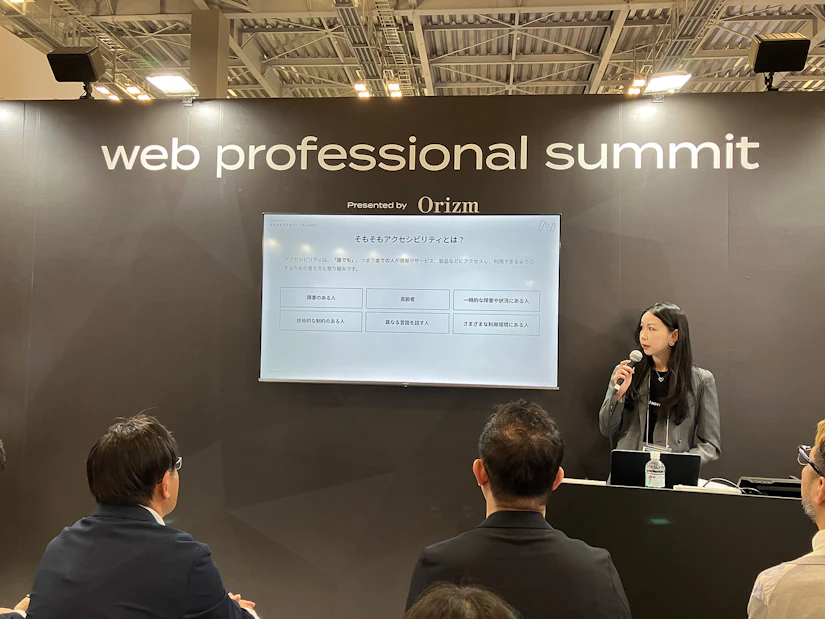 「web professional summit」と書かれたブースの中で、ノベルティの社長がマイクを持って立ち、ノベルティのアクセシビリティへの取り組みについての話をしている。