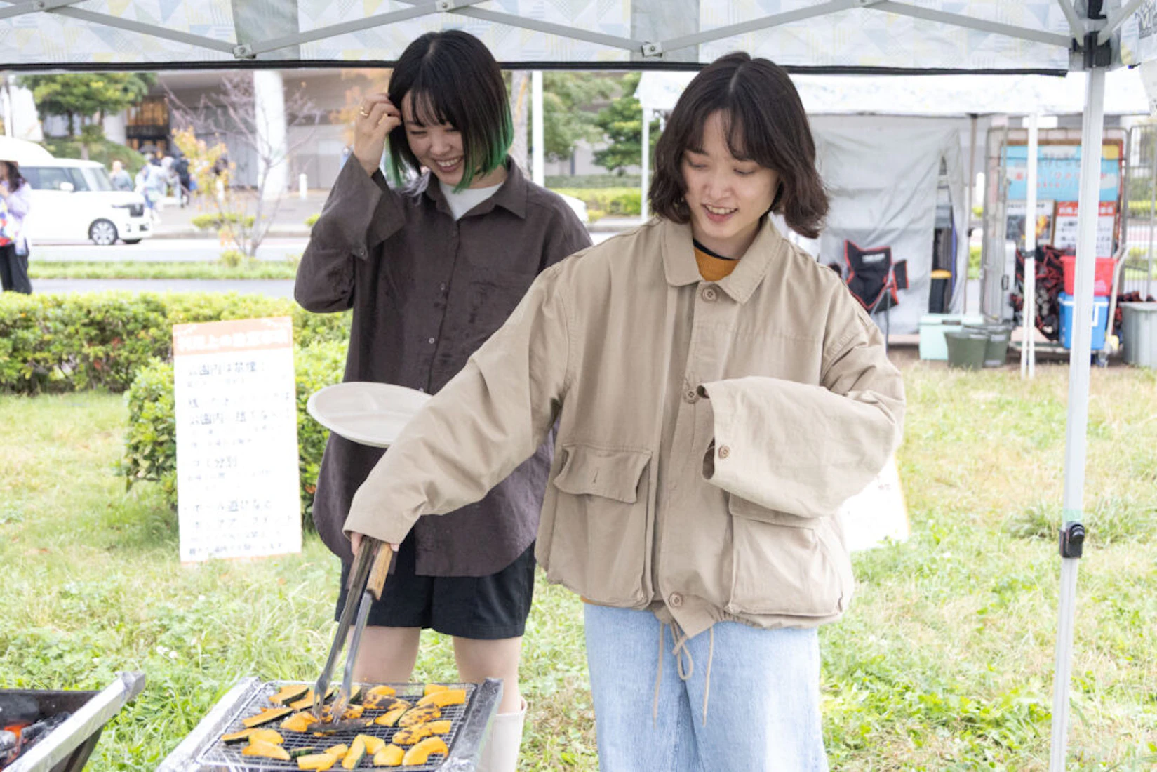 少し怖がりながらコンロで焼いているカボチャをトングでとる人。その後ろにはお皿を持って微笑んでいる人がいる。