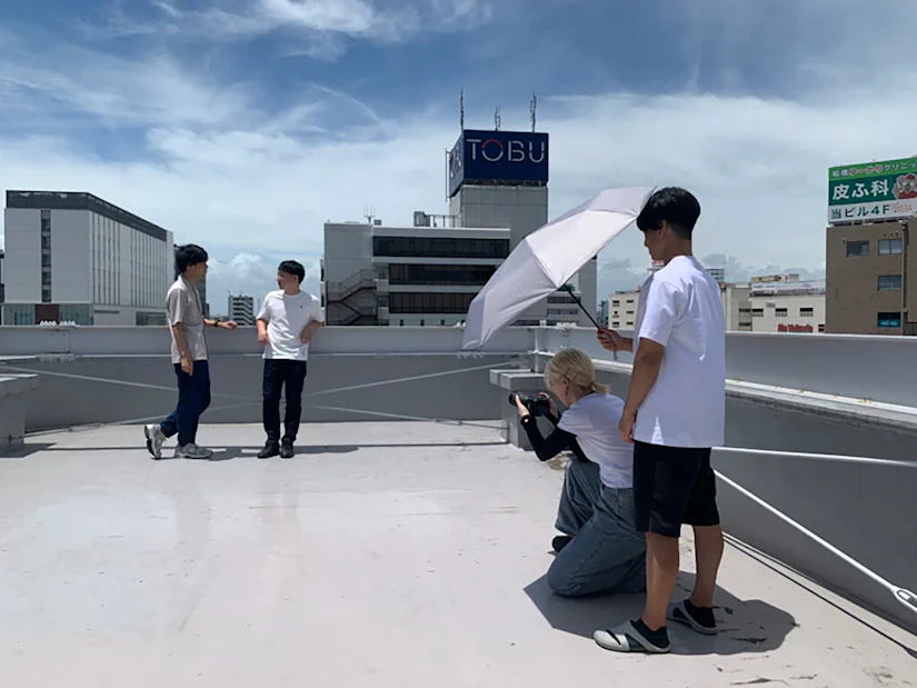 ビルの屋上で撮影会をしている様子。手前にカメラマンがおり、奥に被写体であるノベルティ社員がポーズをとっている。