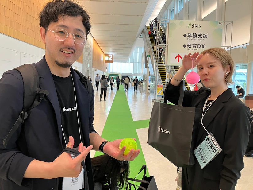 ノベルティ社員2名が、ブースでもらったピンクと黄色のふわふわの毛が生えたヨーヨーを持っている。