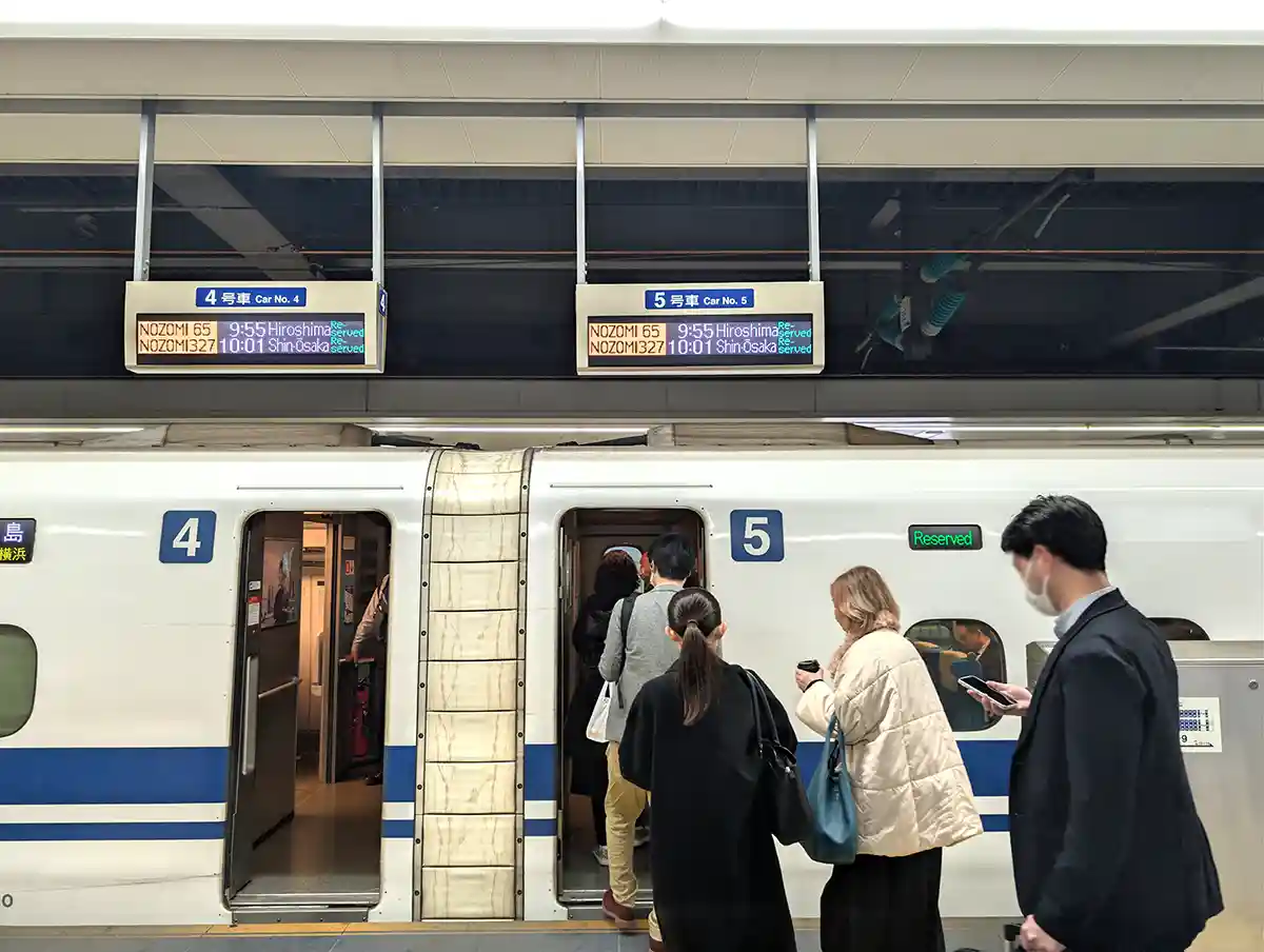 Passengers boarding a Shinkansen train at Shinagawa Station with departure board and car numbers above