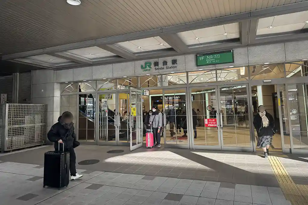 Ground level west exit at Sendai Station with station name signs and people arriving with luggage.