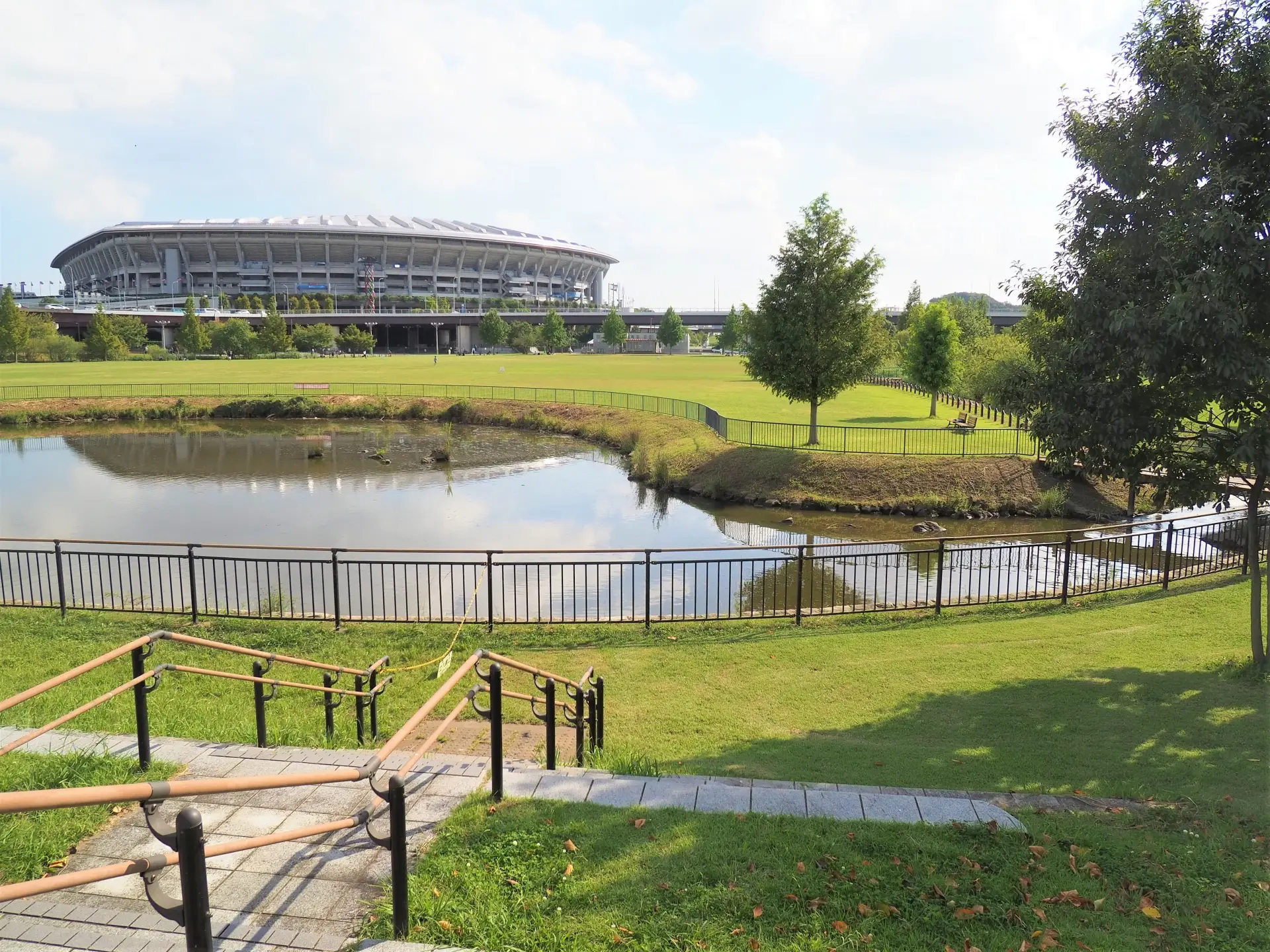 Park area with pond and trees at Shin‑Yokohama Park, with Nissan Stadium visible in the background