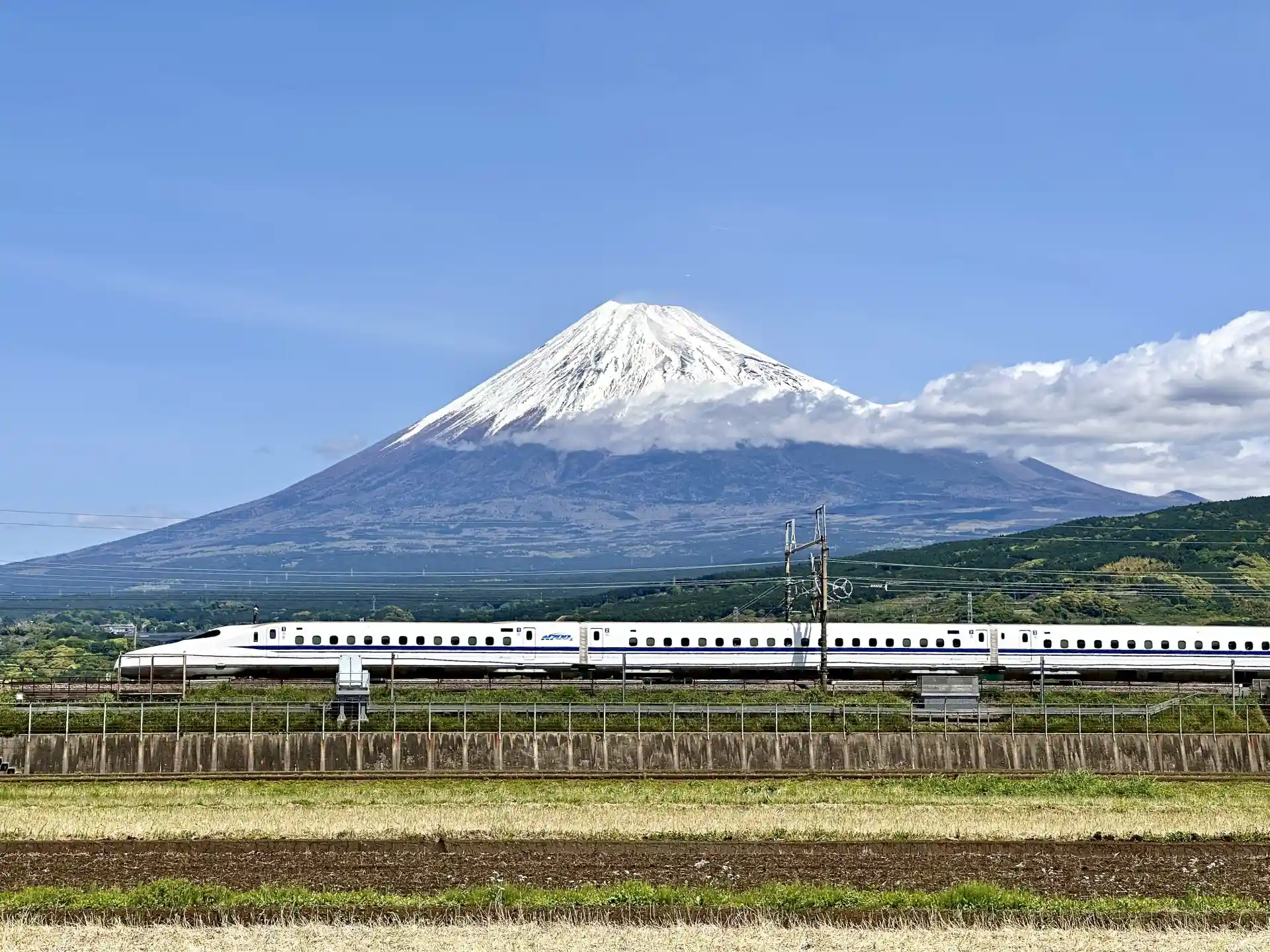 A Tokaido Shinkansen train passing in front of Mount Fuji, showing the snow-covered peak and wide rural landscape under a clear sky