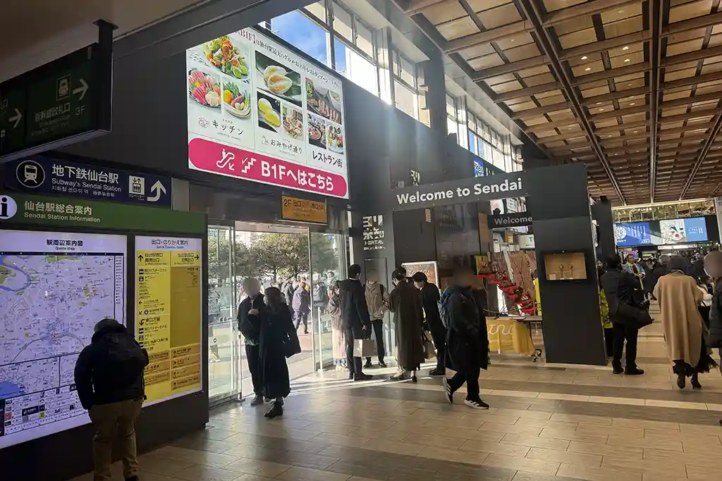 West exit area of Sendai Station on the 2F level where passengers arrive by escalator and gather near information signs.