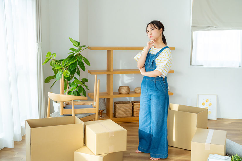 家で多くのダンボールを広げ、小首を傾げて悩んだ表情を浮かべる女性。