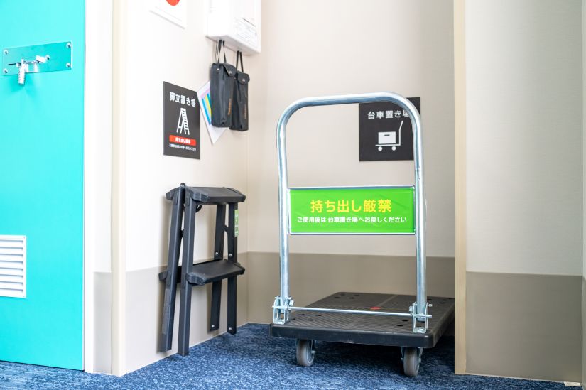 ストレージ王亀戸トランクルームのエントランスに用意された、荷物運びに便利な大きめの台車と、高い場所への荷物の出し入れに役立つ脚立の画像。