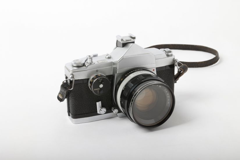 銀色のボディに黒い革張りの部分、レンズが取り付けられたフィルム一眼レフカメラ。上部にはアクセサリーシューと巻き上げレバー、シャッターボタンが見える。革製のストラップが右側に取り付けられている。
