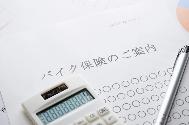 バイク保険のご案内と記載された書類の上に、電卓とボールペンが置かれている