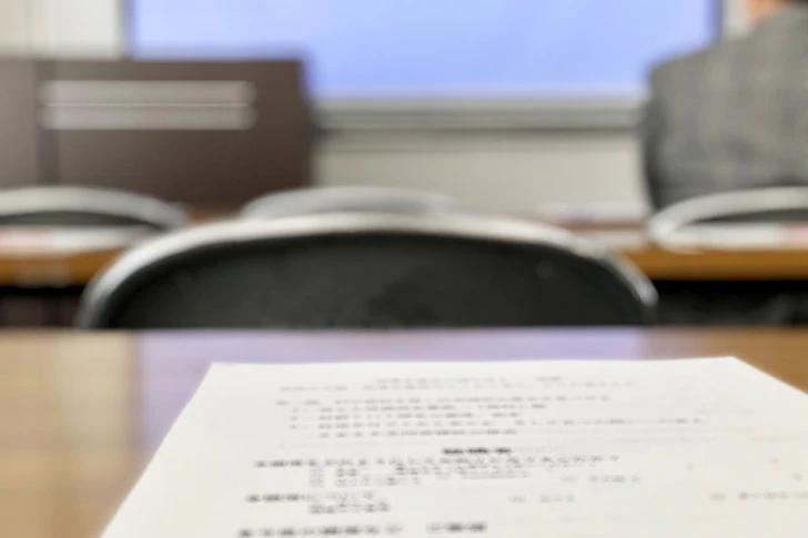 手前のデスクに置かれた試験問題用紙と、奥に見える試験会場の様子。