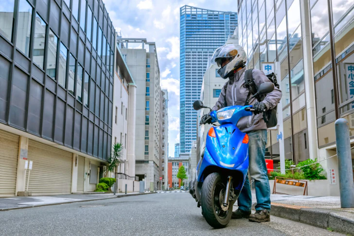青いバイクから降りて支えている人、奥にはビルがいくつか見える