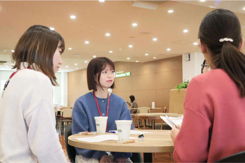 明るいカフェテリアで3人の若い女性が丸テーブルを囲んで立って話をしている様子。1人は青いセーター、1人は白いセーター、1人は赤いトップスを着ている。テーブルの上には飲み物の入ったカップがある。